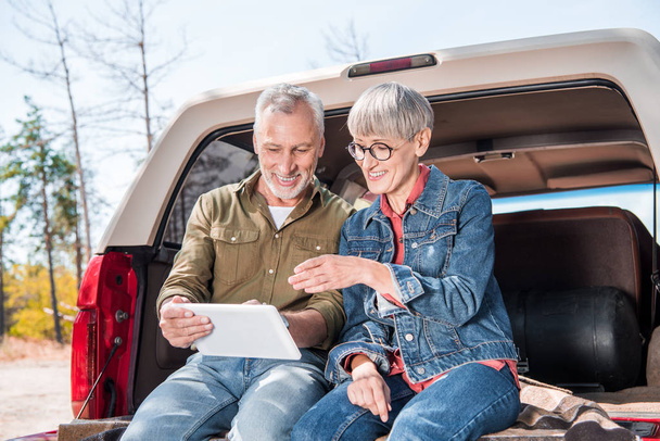 Lächelndes Senioren-Paar sitzt im Auto und nutzt an sonnigen Tagen digitales Tablet - Foto, Bild