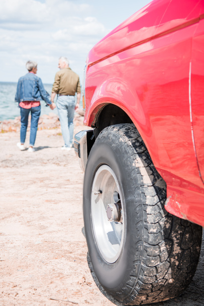 Вид в полный рост пожилой пары, держащейся за руки во время прогулки по пляжу, и красный автомобиль на переднем плане
 - Фото, изображение