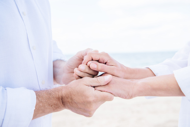 cropped view of senior couple in white shirts holding hands under blue sky - Photo, Image