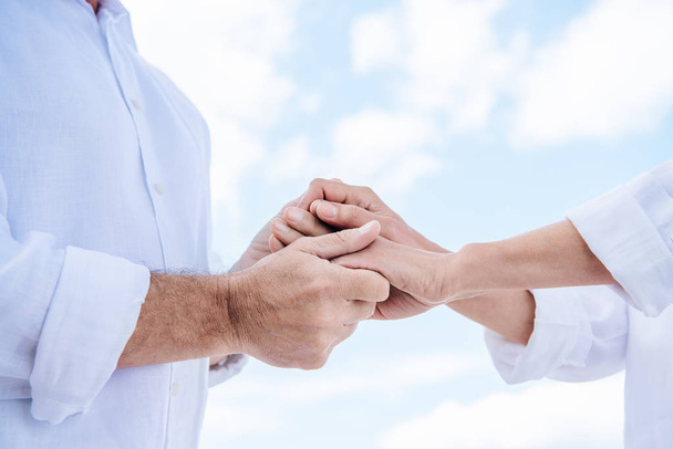 cropped view of senior couple in white shirts holding hands under blue sky - Photo, Image