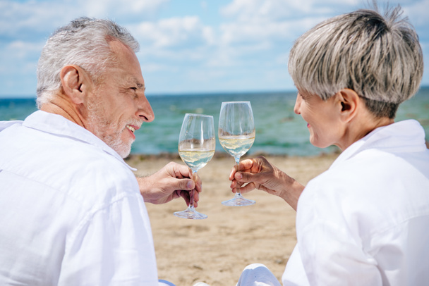 hymyilevä vanhempi pari pitelee viinilaseja viinillä ja katselee toisiaan rannalla
 - Valokuva, kuva
