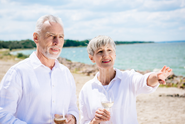 smiling senior couple holding wine glasses with wine and looking away at beach - Fotoğraf, Görsel