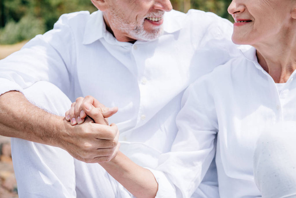 cropped view of smiling senior couple in white shirts holding hands - Valokuva, kuva