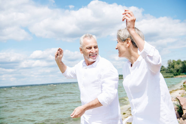 feliz sonriente pareja mayor en camisas blancas mirándose unos a otros y haciendo gestos en la playa bajo el cielo azul
 - Foto, Imagen