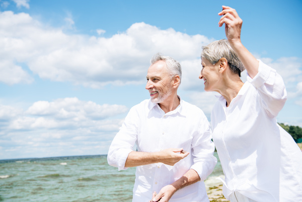 happy smiling senior couple in white shirts gesturing at beach under blue sky - Fotoğraf, Görsel