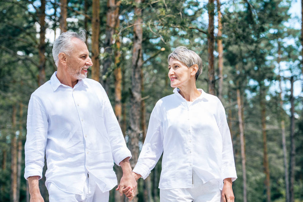 Gelukkig Senior paar in witte shirts vasthouden van handen en kijken naar elkaar in bos - Foto, afbeelding