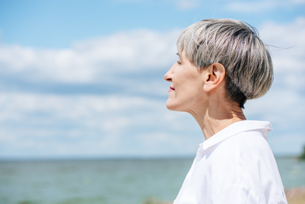 πλευρική όψη της ανώτερης γυναίκας με λευκό πουκάμισο κοιτάζοντας μακριά κοντά στο ποτάμι την ηλιόλουστη μέρα - Φωτογραφία, εικόνα