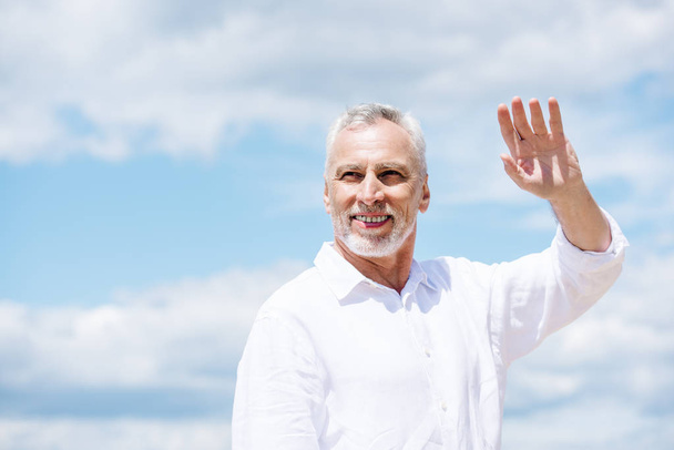glimlachend Senior man met baard op zoek weg en zwaaiende hand onder de blauwe hemel in zonnige dag - Foto, afbeelding