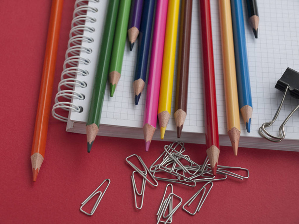 Material escolar e de escritório: lápis de cor, clipes de papel, ligantes e caderno sobre um fundo de papel vermelho. Vista superior. Close-up
 - Foto, Imagem