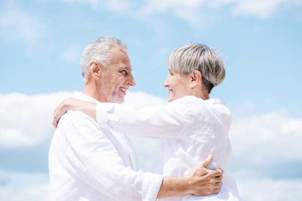 vista lateral de la sonriente pareja de ancianos abrazándose y mirándose bajo el cielo azul
 - Foto, imagen