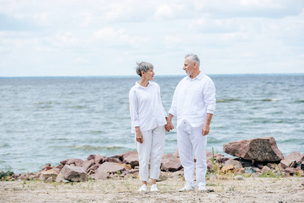Ganzkörperansicht eines älteren Paares, das sich am Strand an den Händen hält und einander ansieht - Foto, Bild