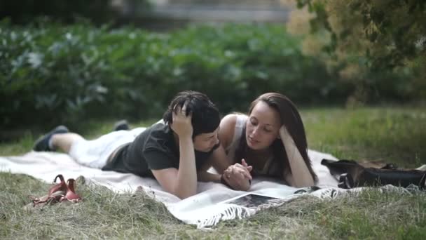 video, Two beautiful lesbians laying on blanket on green grass in park. Lesbians holding hands. The girls are kissing in lips and smiling - Video, Çekim
