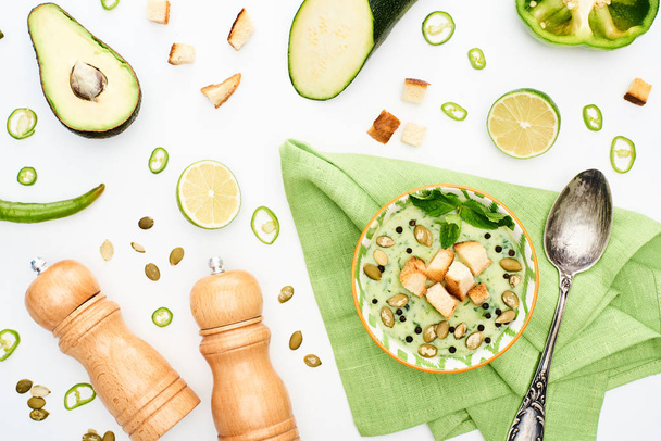 вид на вкусный сливочно-зеленый овощной суп рядом с перцово-солевыми мельницами и разбросанными ингредиентами, изолированными на белом
 - Фото, изображение