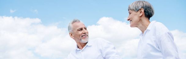 panoramic view of smiling senior couple in white shirts looking at each other under blue sky - Photo, Image