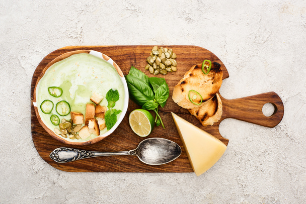 top view of green vegetable creamy soup on wooden chopping board near cheese, croutons and spoon on textured surface - Foto, Imagem