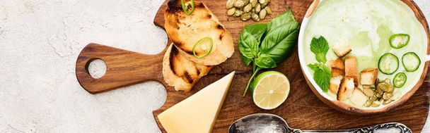 panoráma lövés zöld zöldség tejszínes leves fa vágódeszka közelében sajtot és kanalat textúrázott felület - Fotó, kép