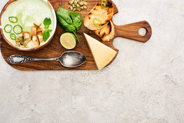 top view of green vegetable creamy soup on wooden chopping board near cheese and spoon on textured surface - Foto, imagen