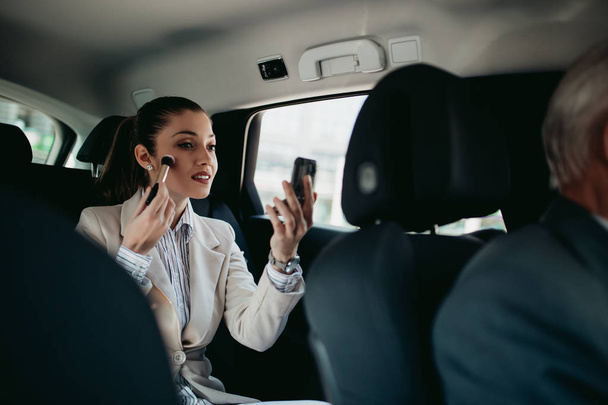 Good looking young business woman sitting on backseat in luxury car and fixing her makeup. Transportation in corporate business concept. - Foto, Bild