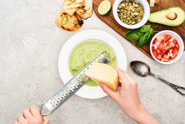 обрезанный вид человека тертый сыр в зеленом сливочном супе возле деревянной доски резки со свежими ингредиентами
 - Фото, изображение