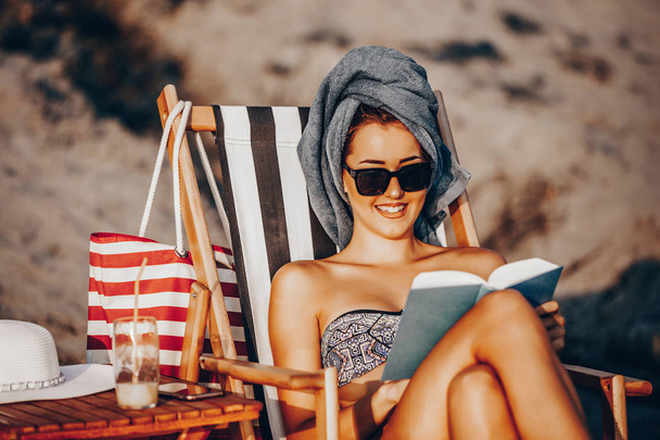 Beautiful young woman in a swimsuit reading a book while sitting - Фото, изображение