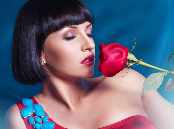 Cute brunette with red rose on blue background in studio - Fotografie, Obrázek