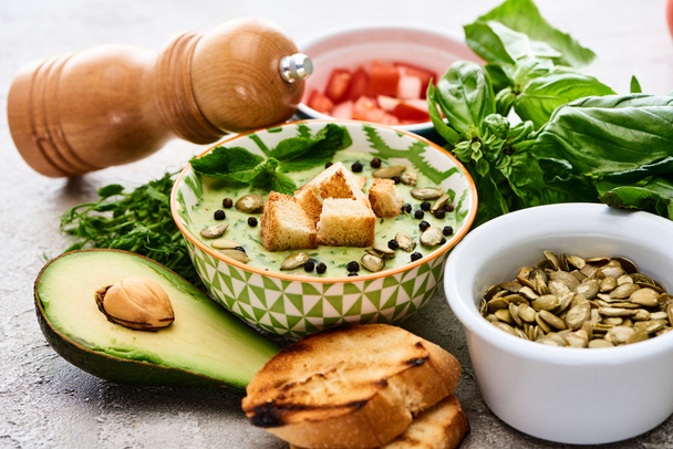 Schüssel mit köstlichem grünen Gemüse cremige Suppe mit Croutons, schwarzem Pfeffer und Kürbiskernen in der Nähe von frischen Zutaten - Foto, Bild