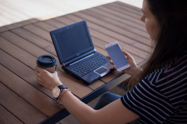 Brunetka dziewczyna z dużym zegarek na rękę, pół-toczone, działa na laptopie stoi na stole. W pobliżu znajduje się smartfon. - Zdjęcie, obraz