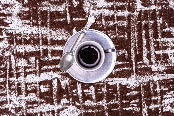 Маленькая чашка кофе и металлическое блюдо с ложкой на стилизованном фоне, образованном узорами вертикальных линий в зернах разбросанного кофе. Вид сверху вниз с графическим фоном, указывающим на чашку
. - Фото, изображение