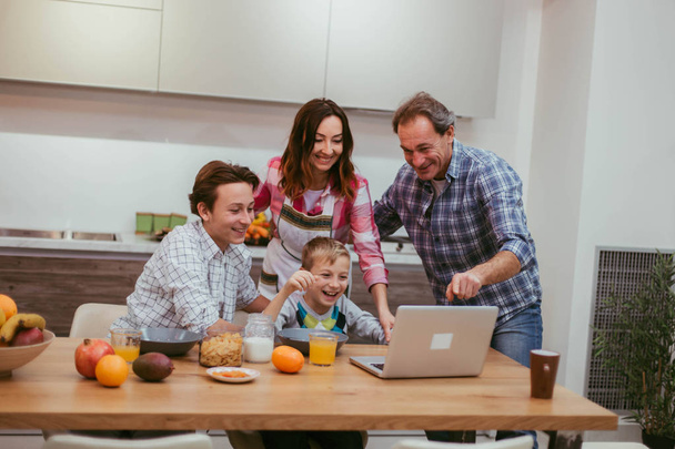 zwei charismatische Jungen mit ihren erwachsenen Eltern beim gemeinsamen Frühstück, Papa zeigt etwas auf seinem Laptop und alle zusammen schauen, modernes Küchendesign im Hintergrund - Foto, Bild