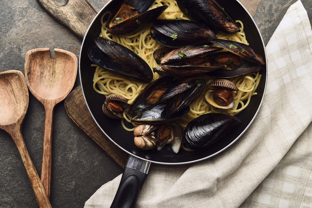 top view of delicious pasta with mollusks and mussels in frying pan on wooden cutting board near napkin and spatulas - Photo, Image