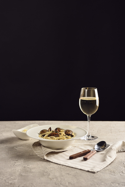 вкусные итальянские спагетти с морепродуктами, подаваемые с белым вином и тертым сыром на салфетке возле столовых приборов, изолированных на черном
 - Фото, изображение