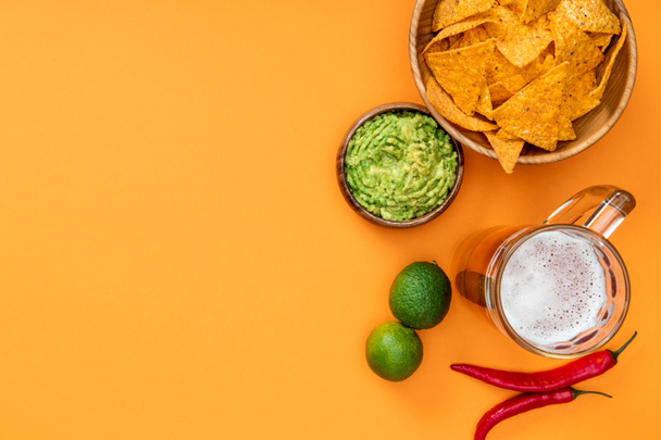 top view of crispy nachos, guacamole, beer, limes and chili peppers on orange background with copy space - Photo, Image