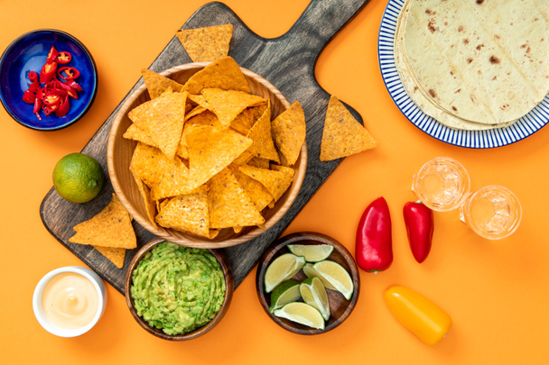 Mexican nachos served on wooden cutting board with guacamole, cheese sauce, peppers, limes, Tequila and tortillas on orange background - Photo, Image