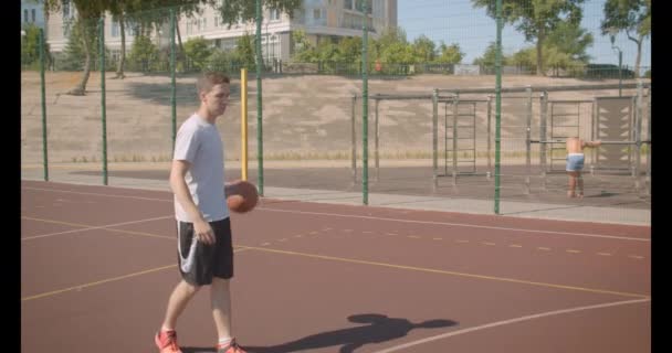 Closeup portrait of young caucasian male basketball player throwing a ball in the hoop on the court outdoors - Metraje, vídeo