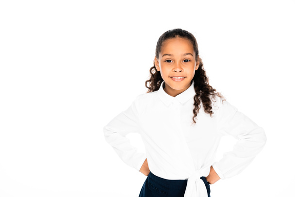 adorable african american schoolgirl smiling at camera while holding hands on hips isolated on white - Фото, зображення