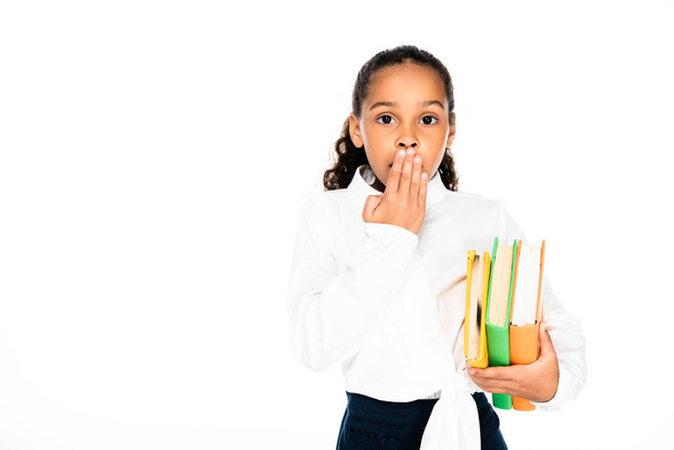 shocked african american schoolgirl covering mouth with hand while holding books isolated on white - Фото, изображение