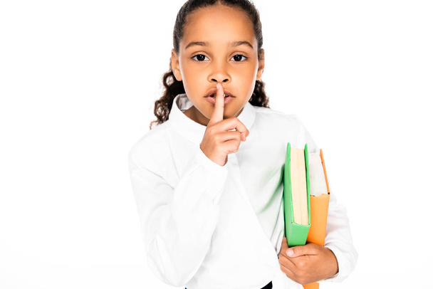cute african american schoolgirl showing hush gesture while holding books isolated on white - Photo, Image