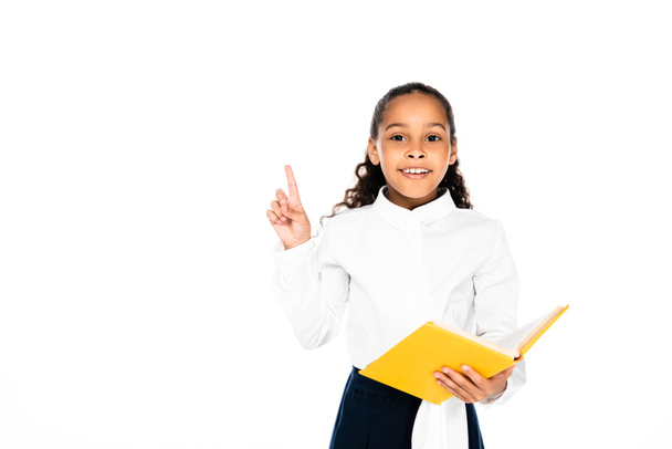 cheerful african american schoolgirl holding book and showing idea gesture isolated on white - Foto, Imagen