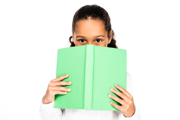 cute african american schoolgirl looking at camera while obscuring face with book isolated on white - Φωτογραφία, εικόνα