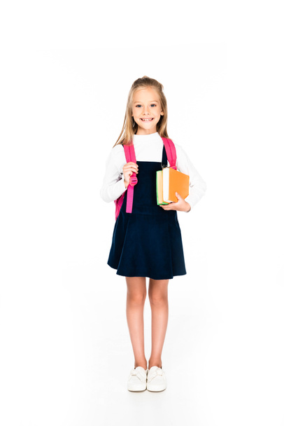 full length view of cute schoolgirl smiling at camera while holding books on white background - Foto, Imagem