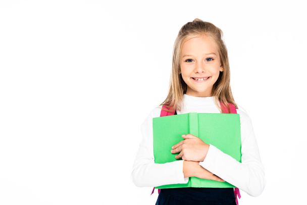 adorable, cheerful schoolgirl holding book and smiling at camera isolated on white - Foto, afbeelding