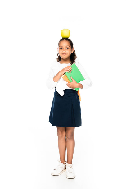 full length view of cheerful african american schoolgirl with apple on head holding books on white background - Foto, Imagen