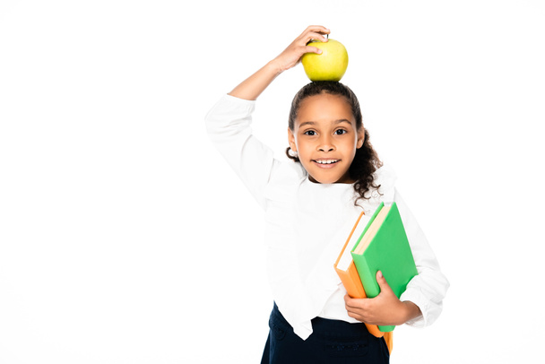 adorable african american schoolgirl holding apple on head while standing with books isolated on white - Fotó, kép