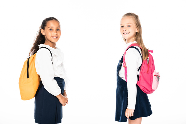 two cheerful multicultural schoolgirls with backpacks looking at camera isolated on white - Fotografie, Obrázek
