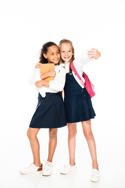 full length view of happy schoolgirl hugging african american friend and taking selfie on white background - Foto, imagen