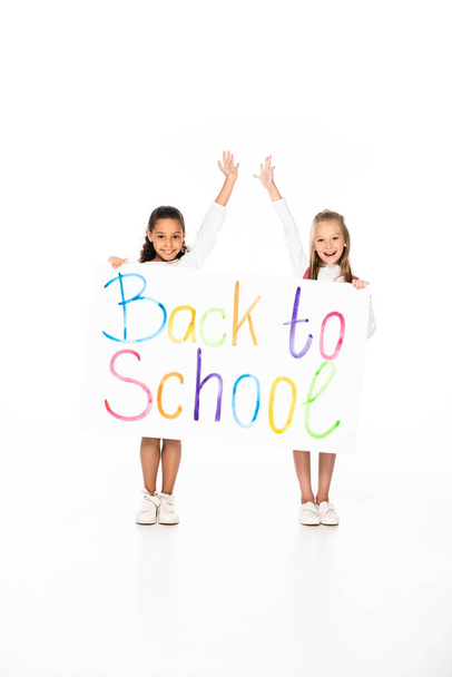 two cheerful multicultural schoolgirls holding placard with back to school inscription on white background - Foto, immagini