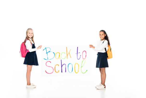 full length view of two multicultural schoolgirls with backpacks holding placard with back to school inscription on white background - Foto, Imagem