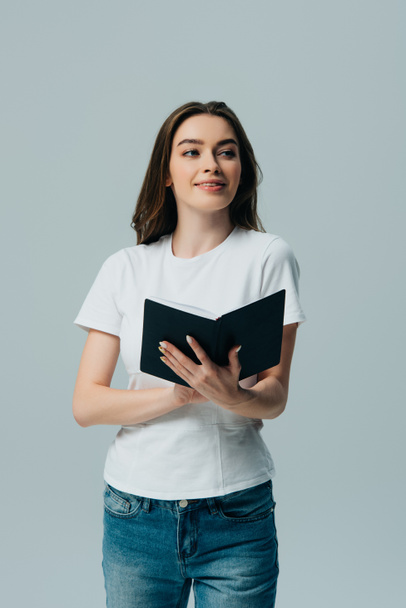 happy beautiful girl in white t-shirt holding notebook and looking away isolated on grey - Фото, изображение