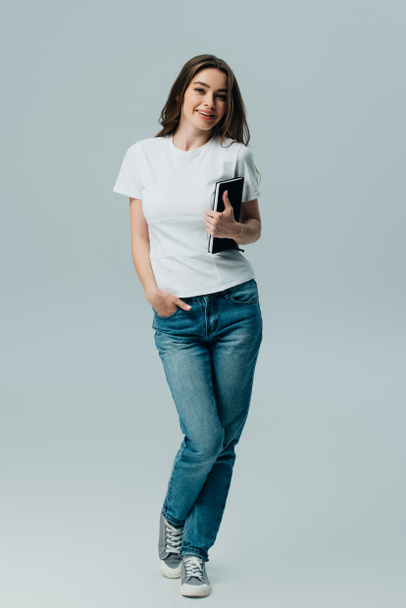 happy beautiful girl in white t-shirt and jeans holding notebook isolated on grey - 写真・画像