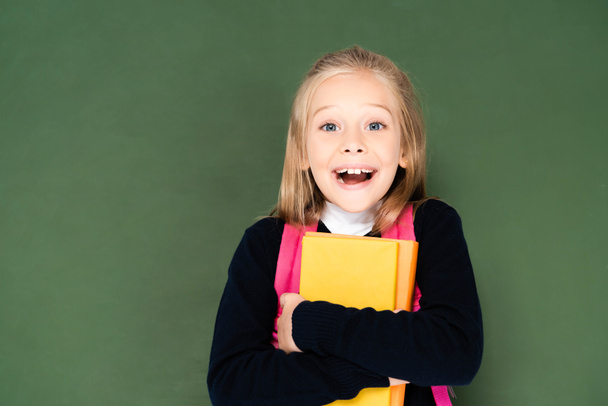 happy schoolgirl looking at camera while holding book and standing near green chalkboard - Photo, Image
